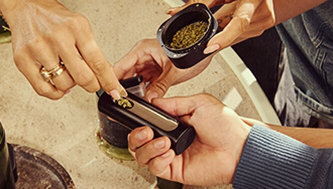 A hand refilling the Pax Flow vaporizer's chamber with fresh ground cannabis using a small scoop from a grinder, set on a marble table for precise reloading.