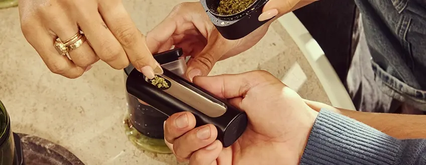 A hand refilling the Pax Flow vaporizer's chamber with fresh ground cannabis using a small scoop from a grinder, set on a marble table for precise reloading.