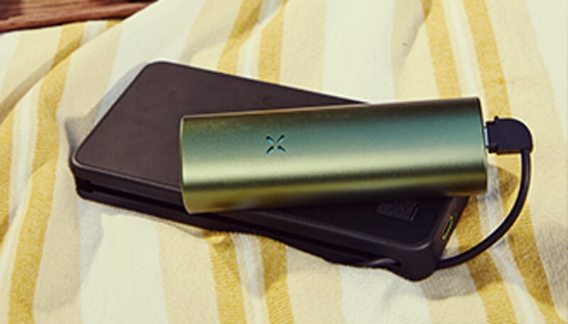 Charging the pax fLow vaporiZer A sleek green pax flow vaporizer connected to a black portable charger via usb cable, resting on a yellow and white striped towel for convenient on-the-go charging.