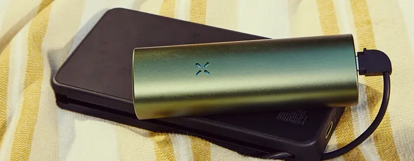 Charging the Pax Flow Vaporizer A sleek green Pax Flow vaporizer connected to a black portable charger via USB cable, resting on a yellow and white striped towel for convenient on-the-go charging.
