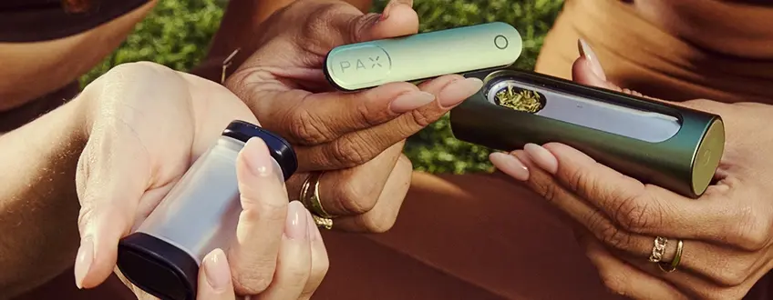 Filling the Pax Flow Vaporizer Two hands collaboratively filling the Pax Flow vaporizer with ground cannabis from a small grinder, showcasing the easy loading process outdoors on green grass.