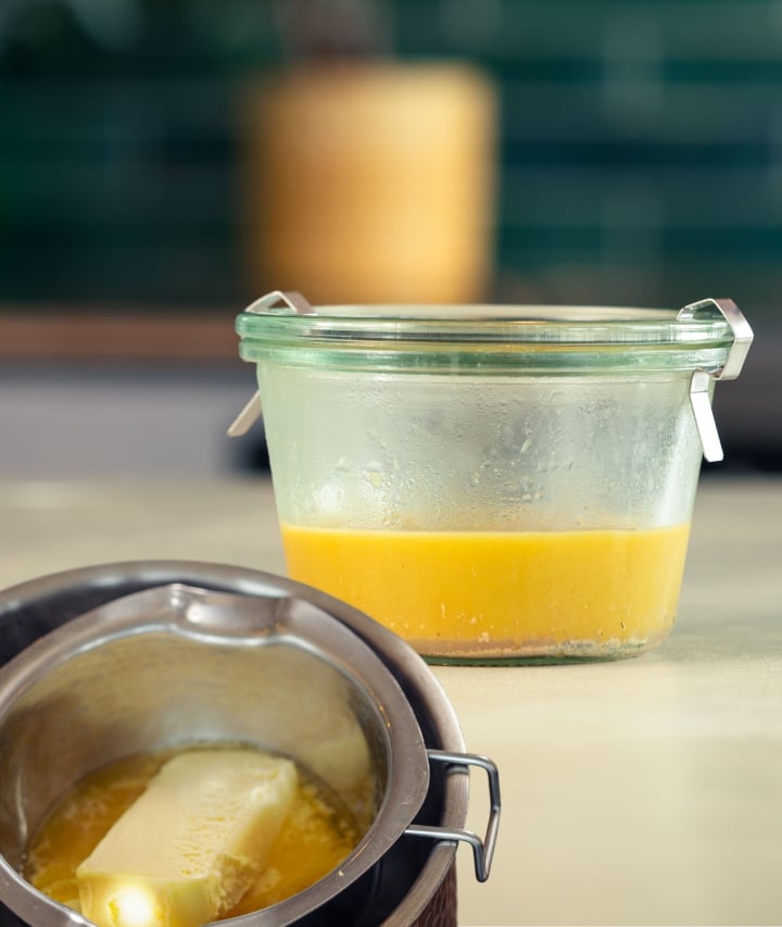 Melted cannabutter being prepared in a pot and transferred into a glass container on a kitchen counter.