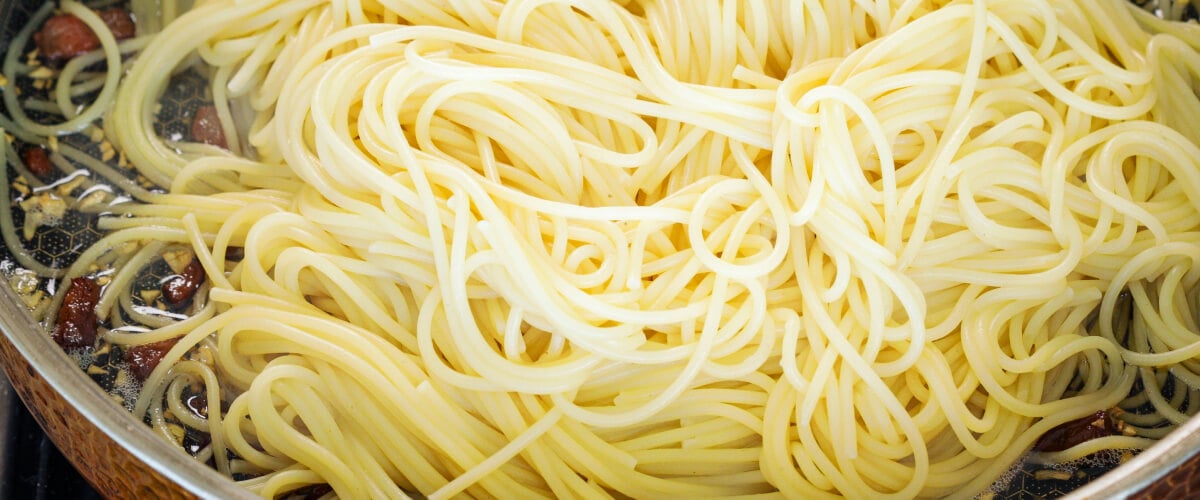 Tossing drained spaghetti with crispy pancetta and infused butter in a pan, fourth step for making cannabis-infused carbonara