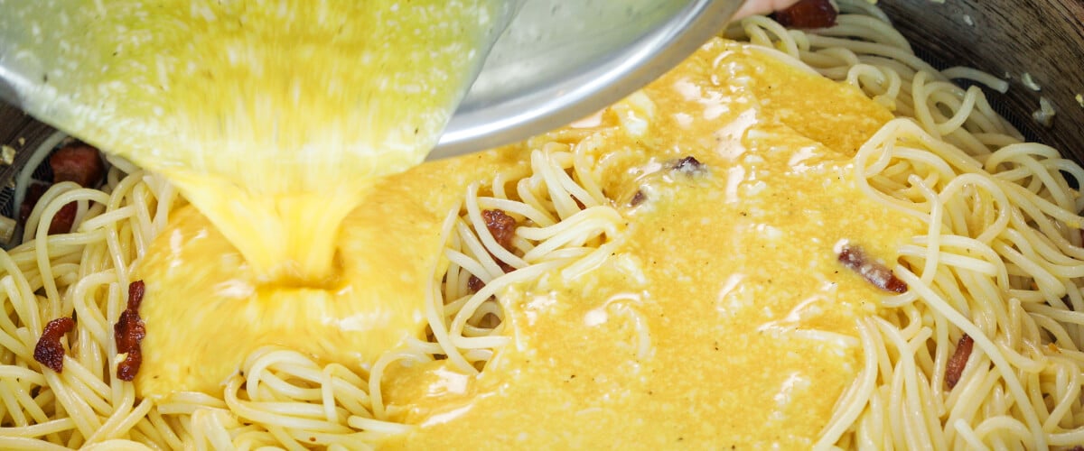 Pouring egg and cheese mixture over hot spaghetti in a pan, fifth step for making cannabis-infused carbonara sauce